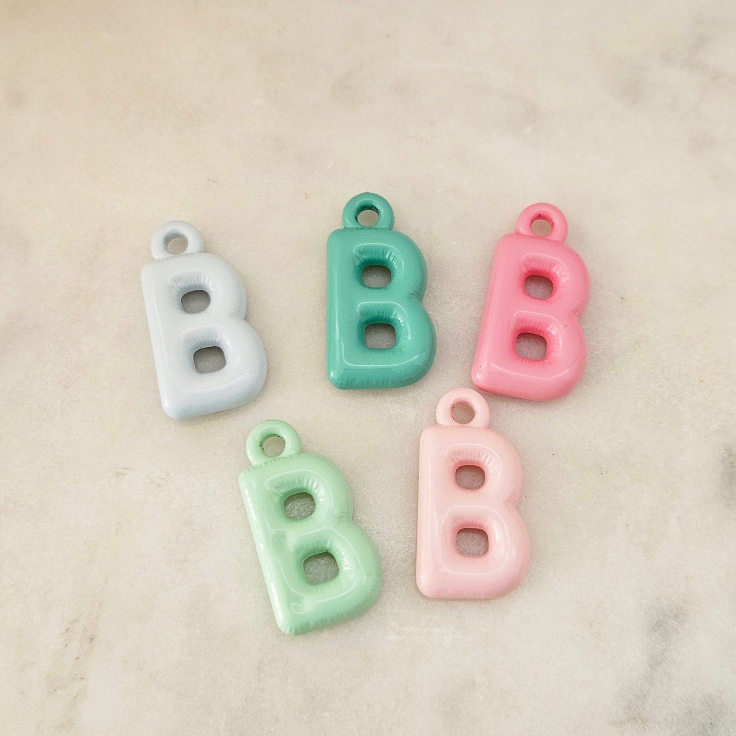 Five pastel letter B charms with hanging loops, shown in white, mint green, pink, and light aqua on a light marble surface.