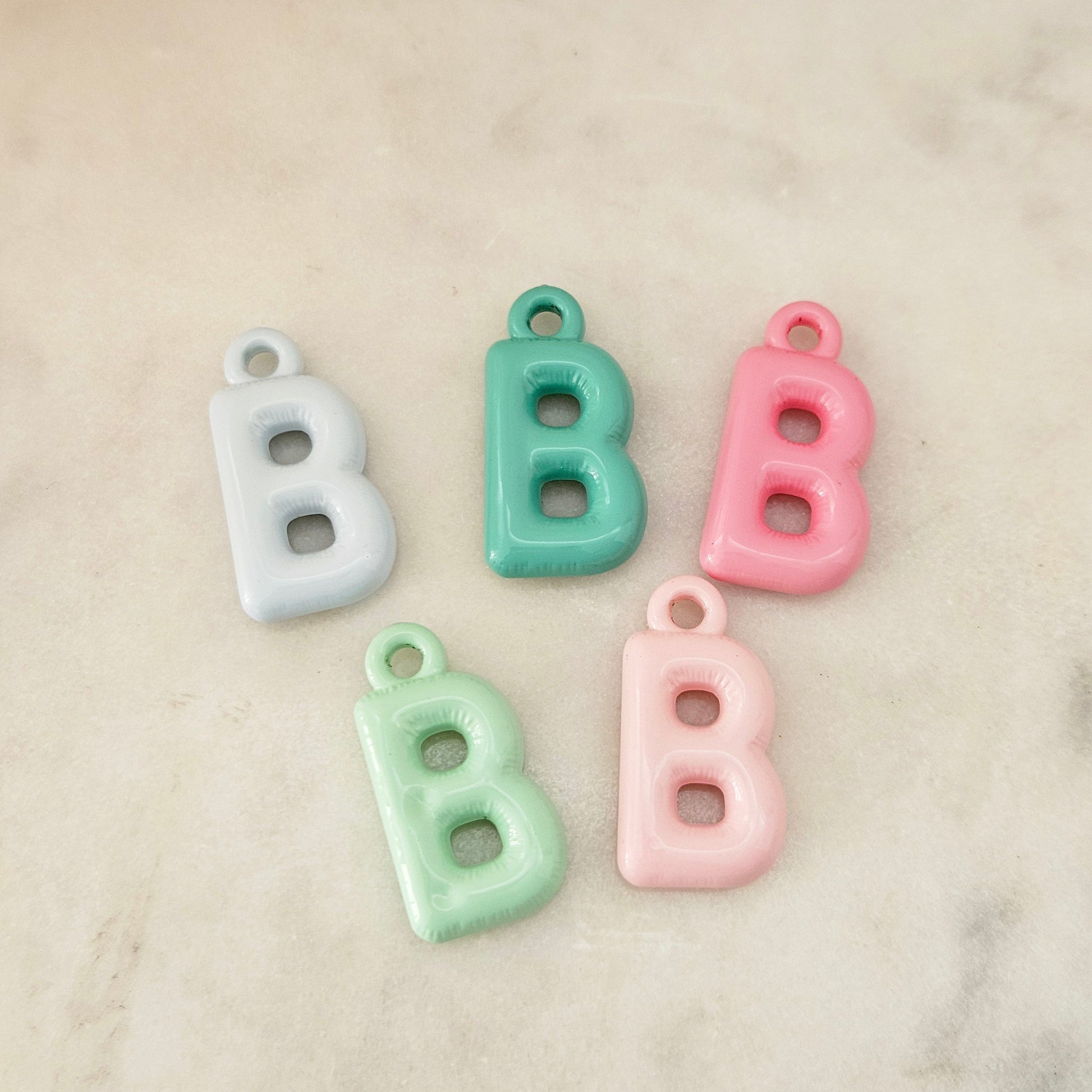 Five pastel letter B charms with hanging loops, shown in white, mint green, pink, and light aqua on a light marble surface.