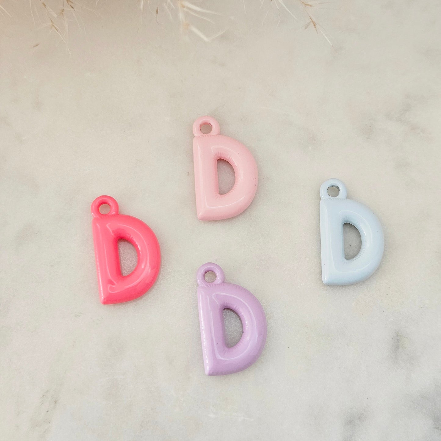 Four glossy letter 'D' charms in pink, light pink, lavender, and light blue, arranged on a light marble surface with dried grass in the top corner.