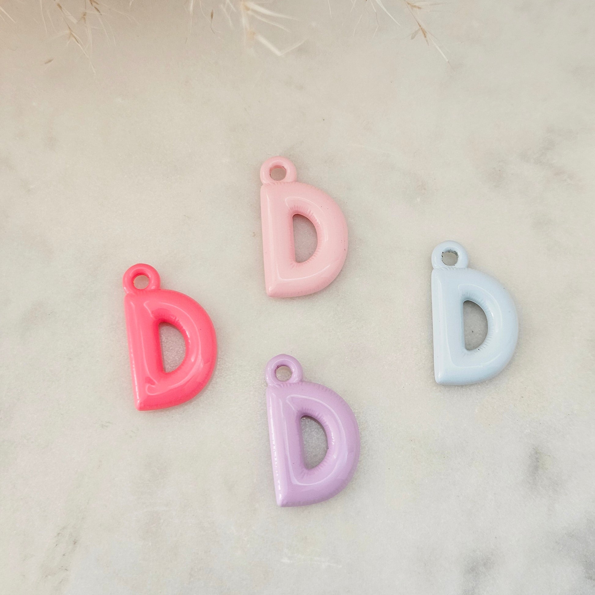 Four glossy letter 'D' charms in pink, light pink, lavender, and light blue, arranged on a light marble surface with dried grass in the top corner.