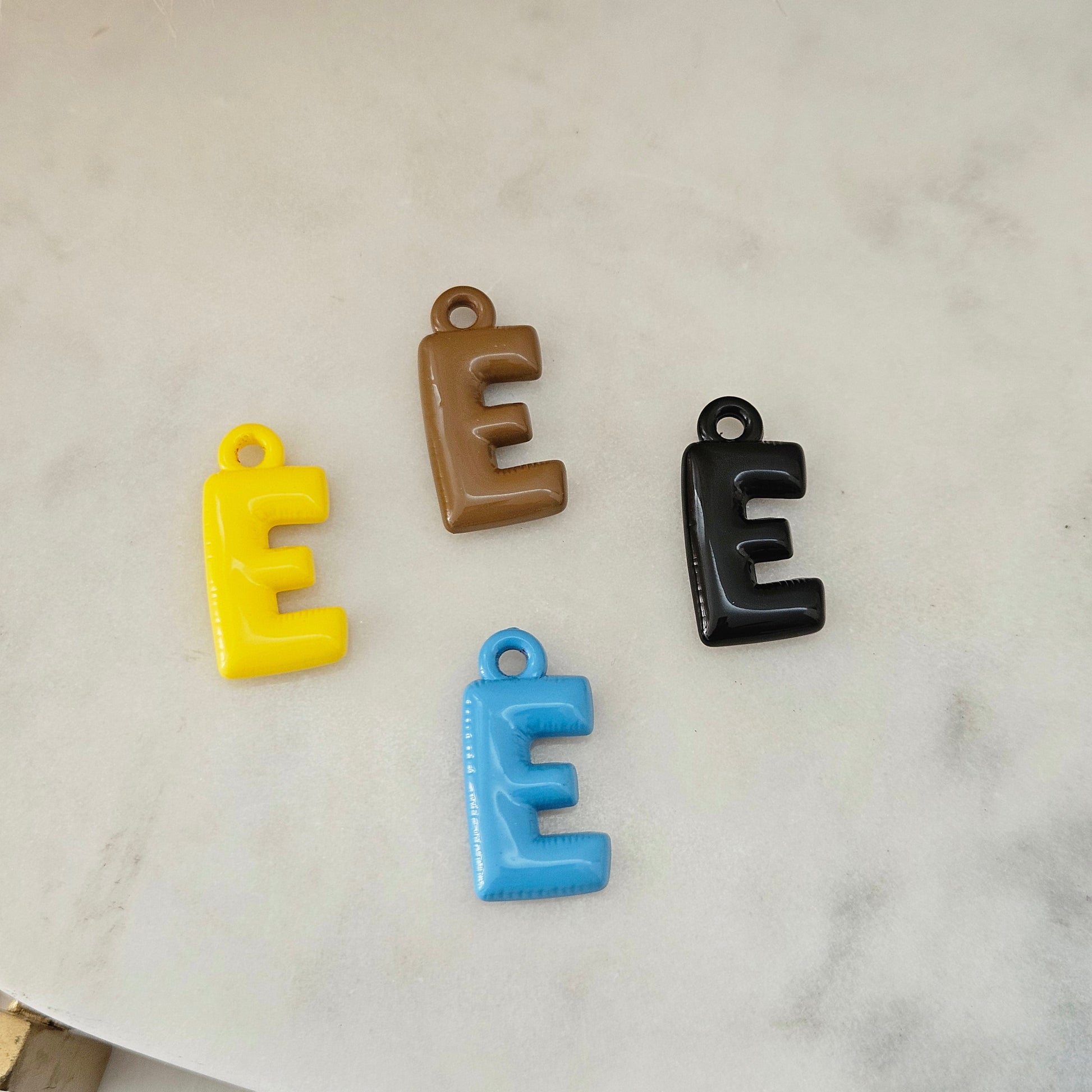 Four small letter E charms in yellow, brown, black, and blue, arranged on a light marble surface.