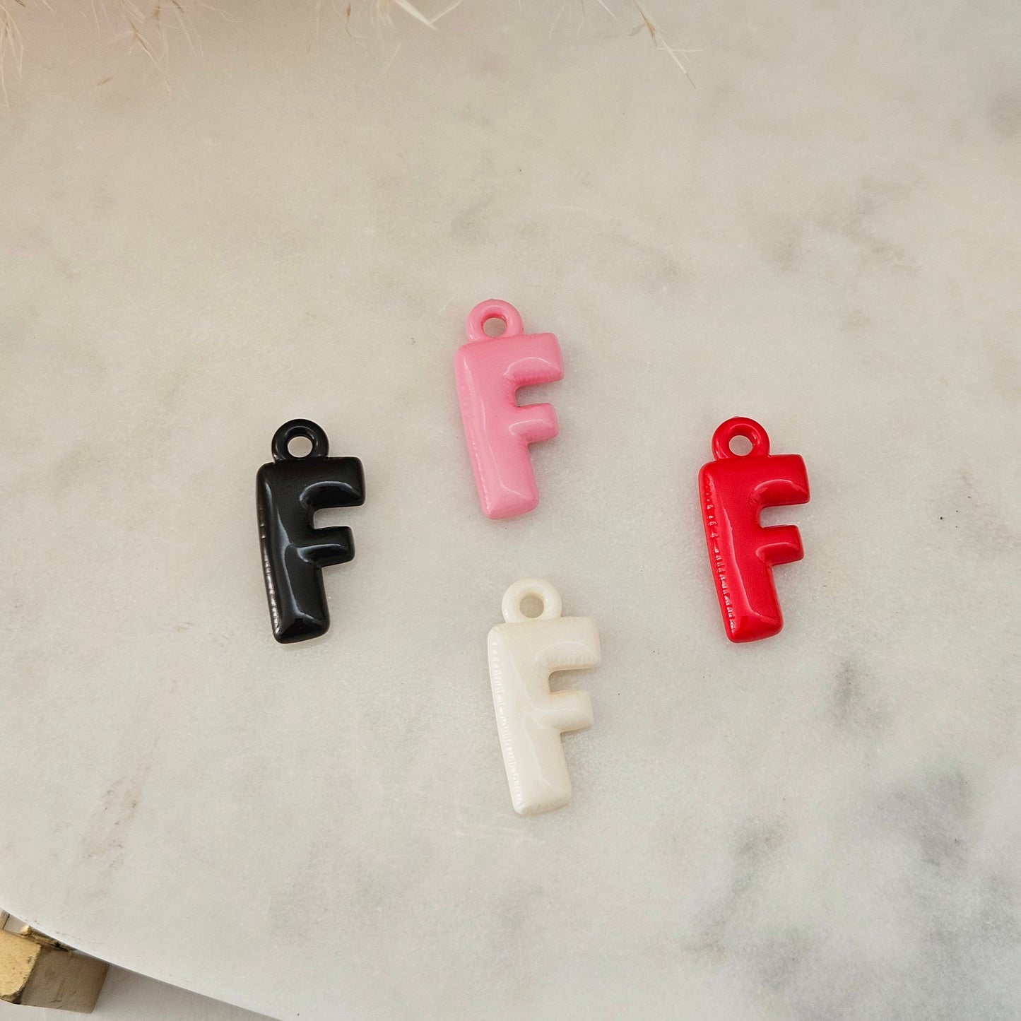 Four small letter F charms in black, pink, red, and off-white, arranged on a light marble surface with dried grass as a decorative prop.