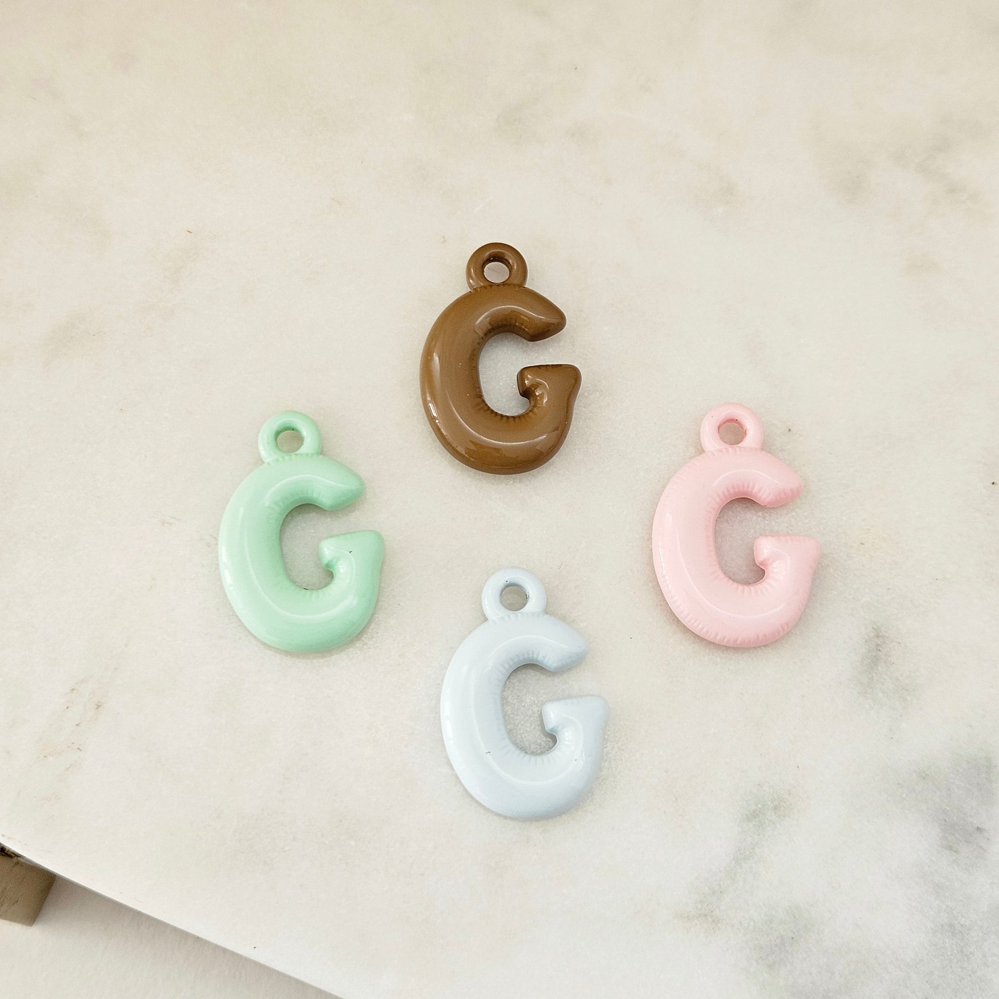 Four glossy letter 'G' charms in mint green, brown, light blue, and light pink, arranged on a light marble surface.