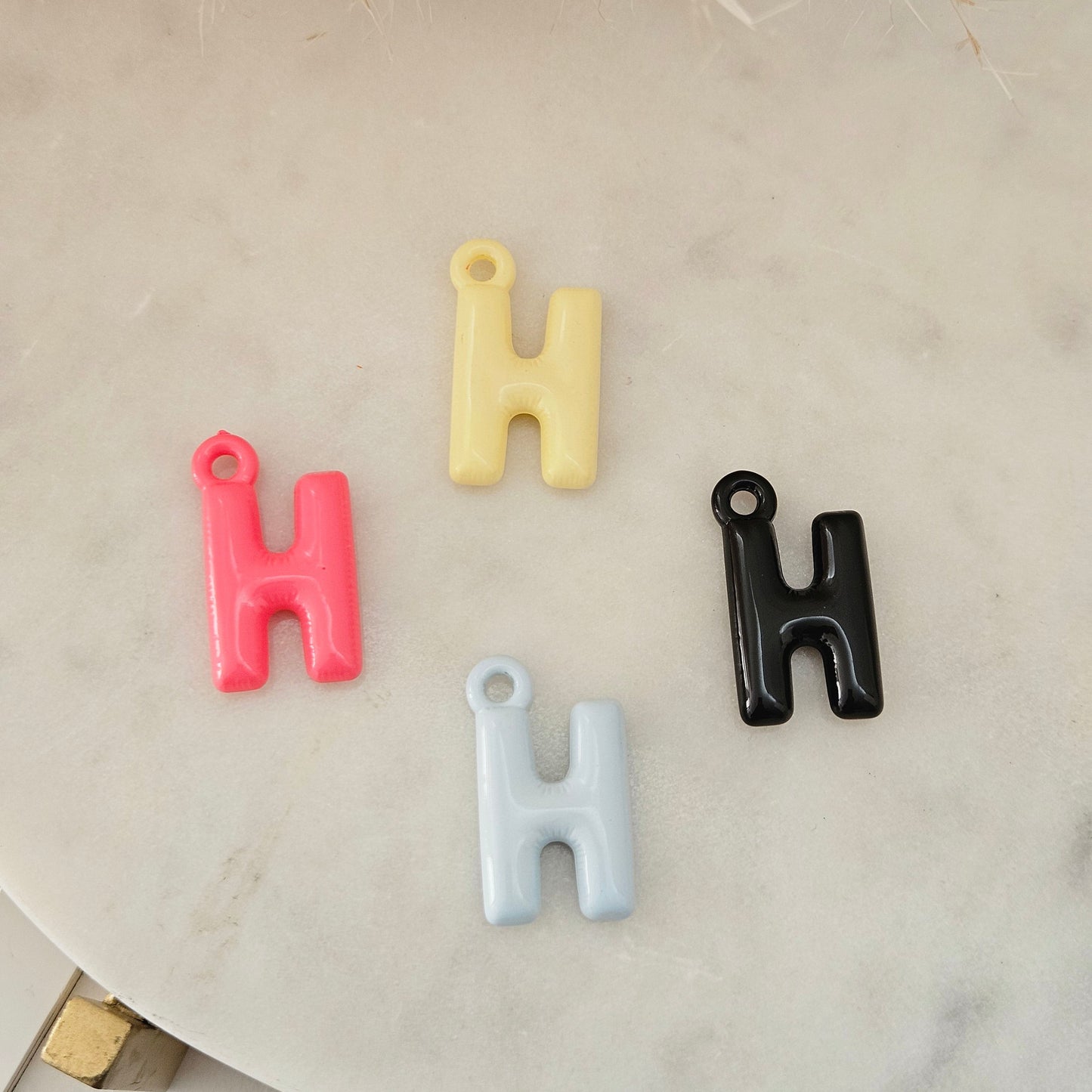 Four small letter H charms in pink, pale yellow, light blue, and glossy black, arranged on a light marble surface with dried grass at the top.