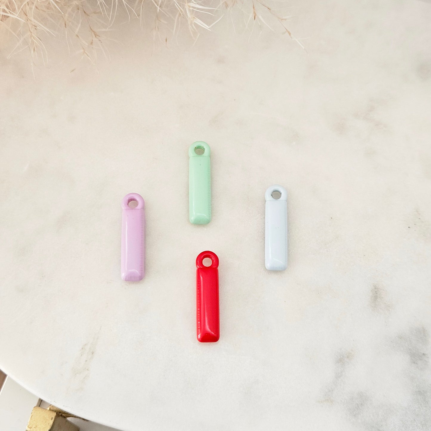 Four small rectangular keychains in pastel purple, mint green, light blue, and bright red, arranged on a light marble surface with dried grass decor in the corner.