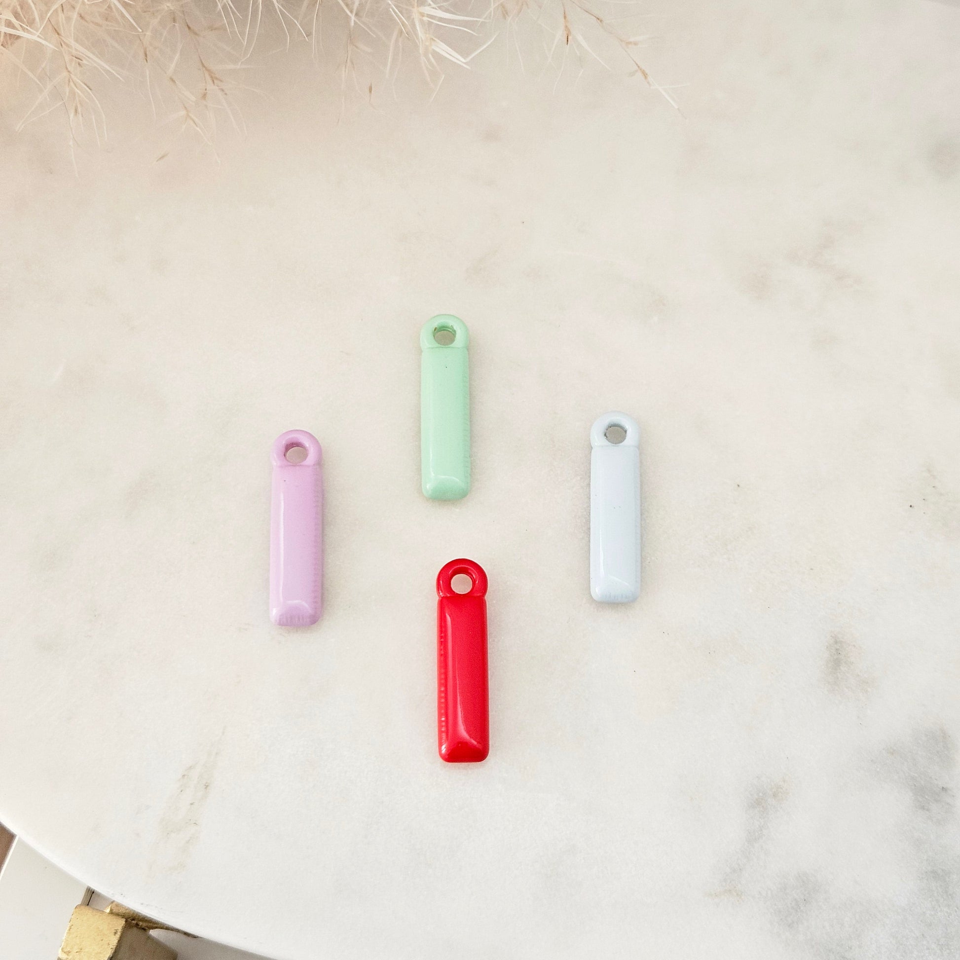 Four small rectangular keychains in pastel purple, mint green, light blue, and bright red, arranged on a light marble surface with dried grass decor in the corner.