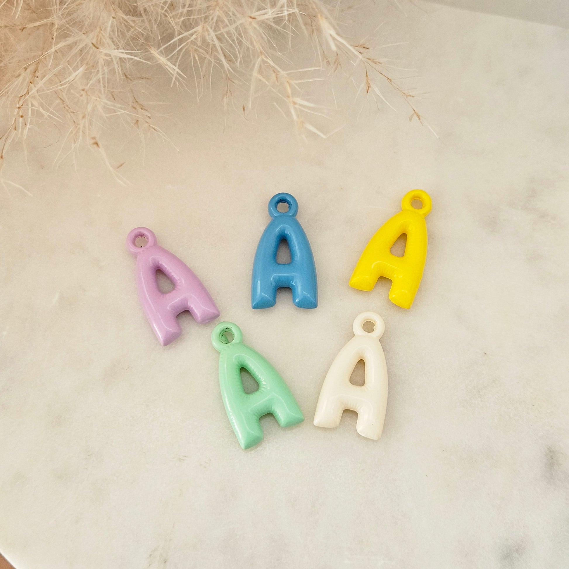 Five colorful letter 'A' charms in pastel shades of purple, blue, yellow, mint green, and cream, arranged on a light marble surface with dried grass in the background.
