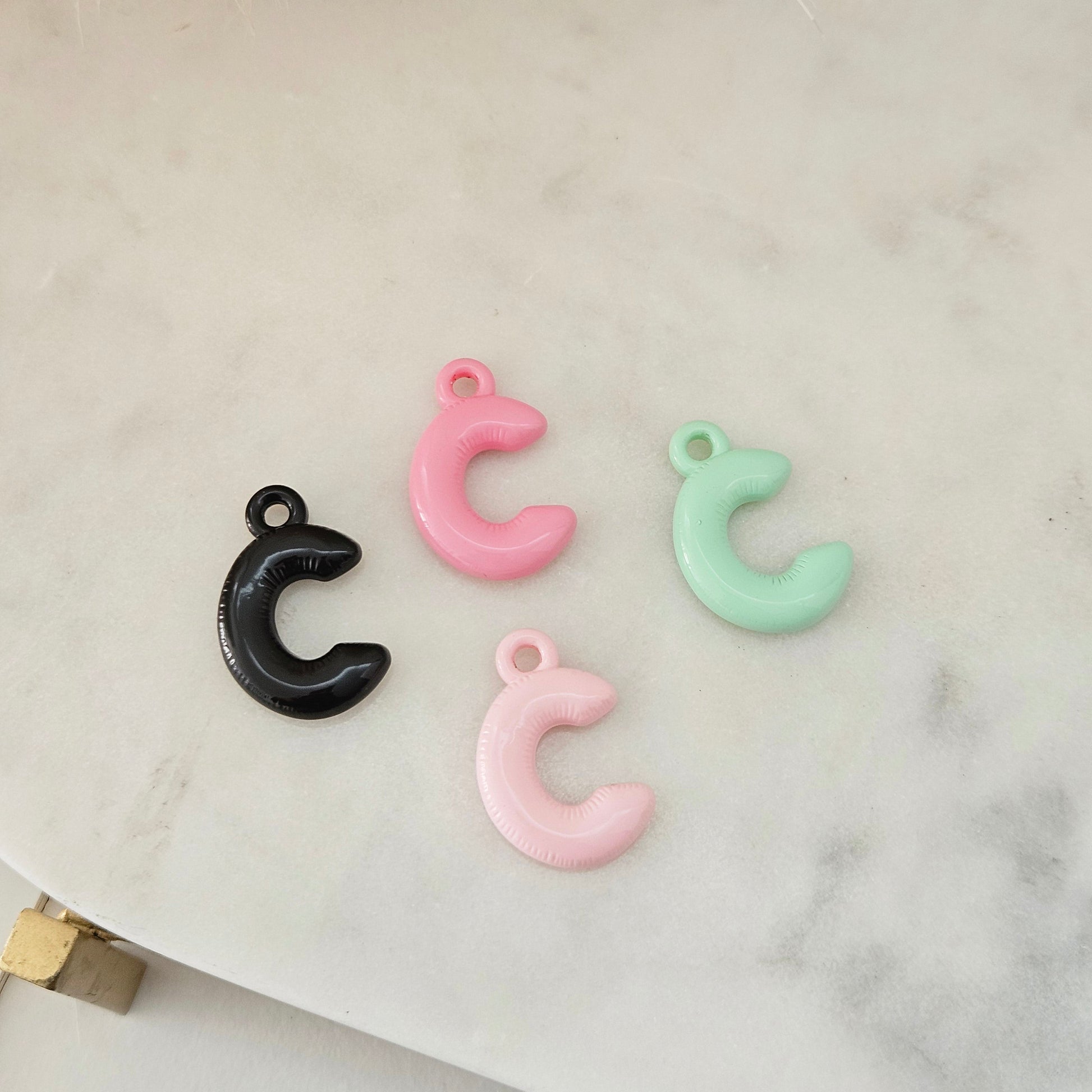 Four small crescent-shaped pendant charms in black, pink, and mint green, arranged on a light marble surface.